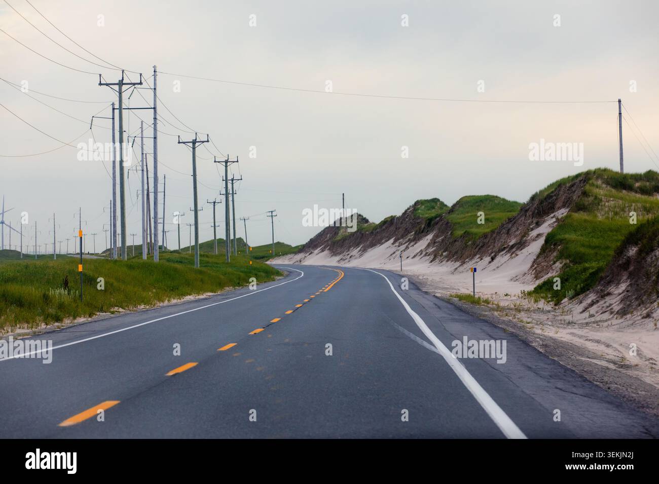 vista di una linea punteggiata gialla curva su una strada asfaltata tra le dune di sabbia e l'erba in una giornata nuvolosa Foto Stock
