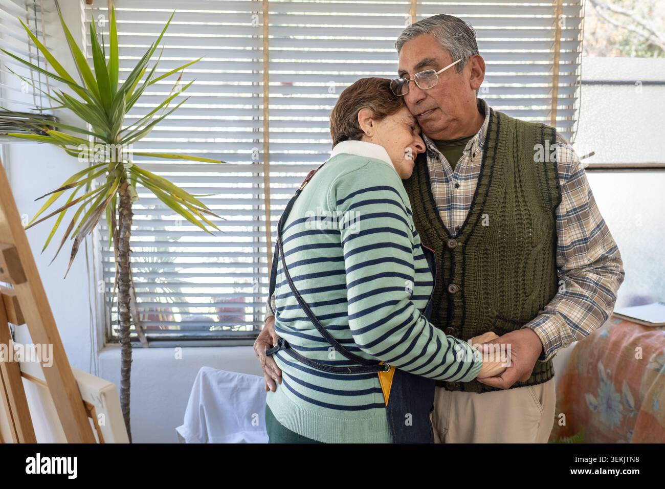 La coppia più anziana condivide un dolce abbraccio simile a una danza vicino a un cavalletto in un soleggiato studio domestico, con le mani strette e i volti vicini in un momento romantico. Foto Stock