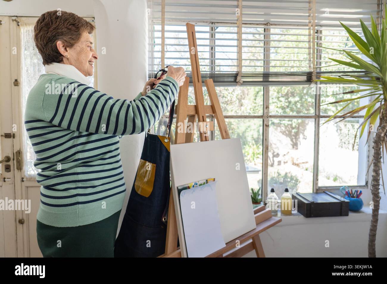 La donna più anziana sorride mentre regola una tela su un cavalletto in legno nel suo luminoso studio domestico, con vista sul giardino attraverso grandi finestre. Foto Stock