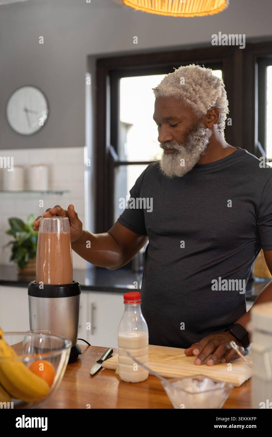 Uomo afroamericano maturo che indossa una maglietta grigia e guarda mentre prepara una bevanda al cioccolato con frullatore e latte Foto Stock