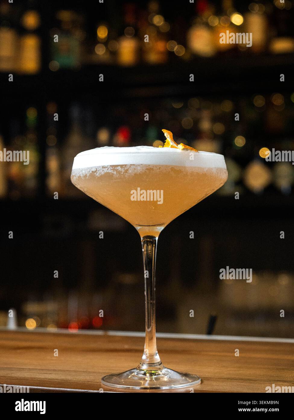 Il cocktail dorato risplende con la schiuma. Lo sfondo delle barre si sfoca, le bottiglie si illuminano con calore. Il vetro poggia su un banco di legno, elegante. Il fumo arricciante aumenta, aggiunge m Foto Stock