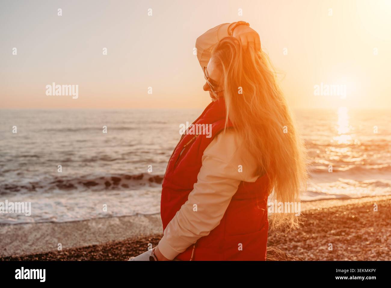 Donna tramonto spiaggia giovane donna bionda in un gilet rosso gode del caldo bagliore del tramonto su una spiaggia di ciottoli in riva al mare Foto Stock