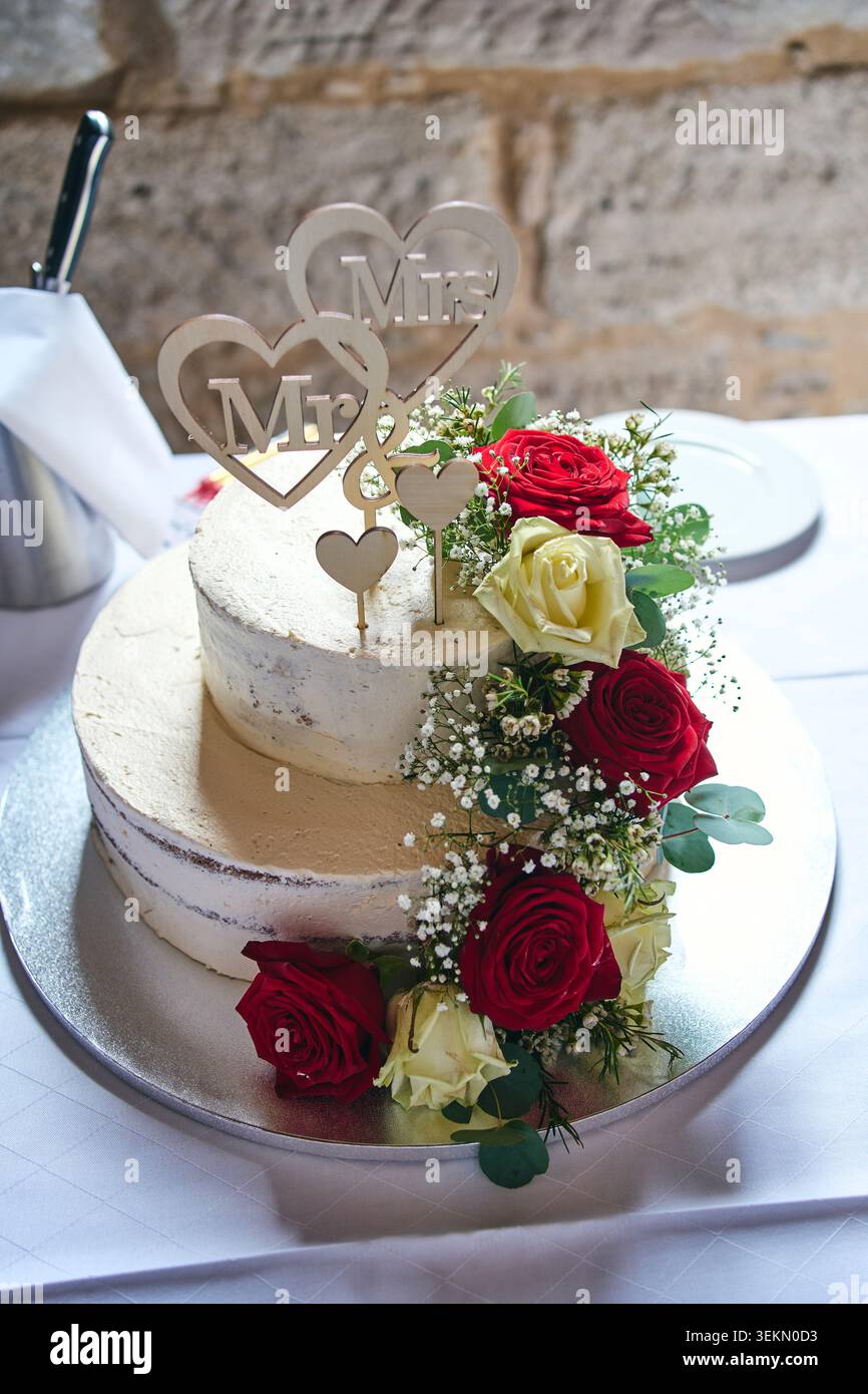 Splendida torta nuziale a due piani decorata con rose rosse e bianche fresche, fiori di respiro per il bambino e un copricuore in legno. Foto Stock