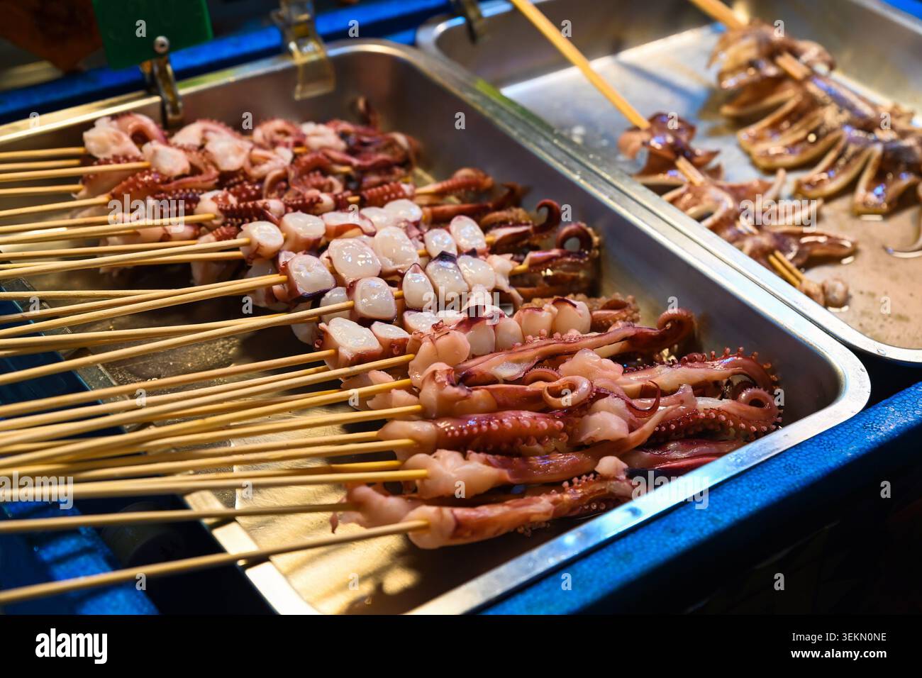 Spiedini freschi di calamari e polpo su vassoi di metallo in vendita al mercato notturno di Temple Street, al chiosco di cibo di strada sotto le calde luci serali di Hong Kong Foto Stock