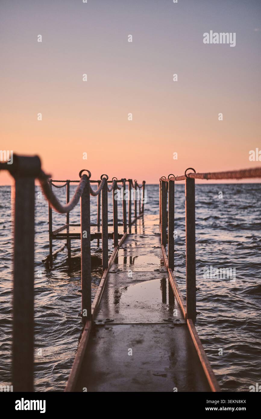 Uno stretto molo in metallo con ringhiere in corda si estende su un'acqua lievemente ondulata verso l'orizzonte, illuminato da caldi colori del tramonto e dalla luce soffusa della sera Foto Stock