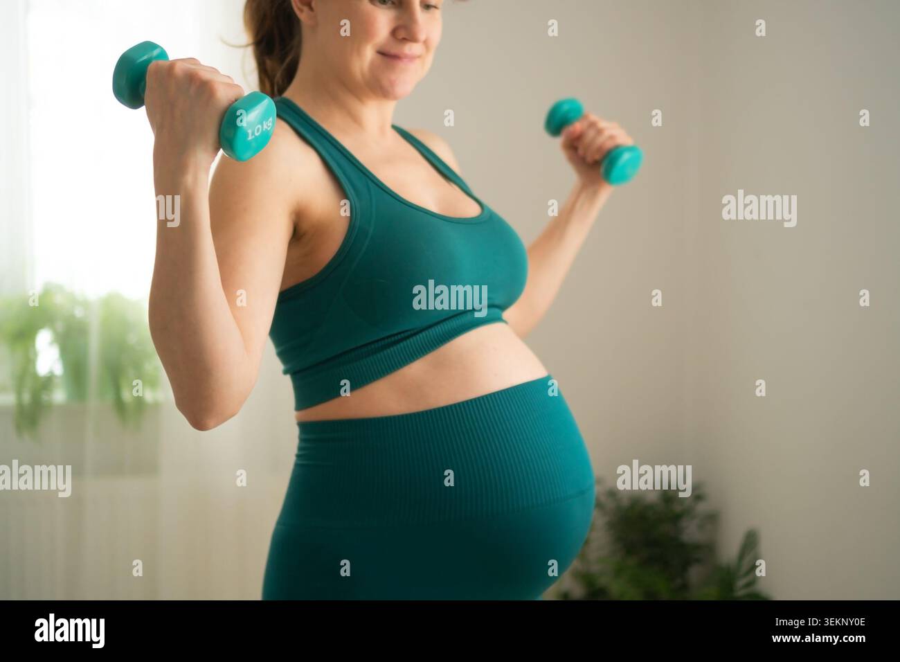 Donna incinta che tiene i manubri in posa sicura per un allenamento in gravidanza in forma. Aspettarsi una madre che rimanga attiva con esercizi leggeri a casa. La futura mamma mostra un forte approccio al fitness prenatale Foto Stock
