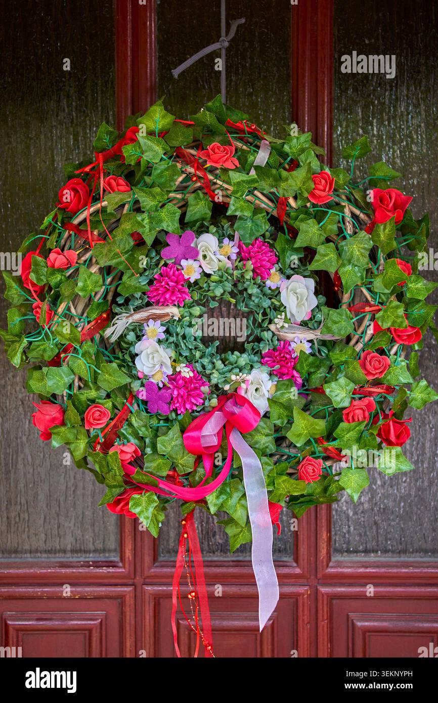 Ghirlanda di porte floreali con foglie di edera verde, rose rosse, fiori rosa e bianchi e nastri colorati appesi su una porta d'ingresso in legno. Foto Stock