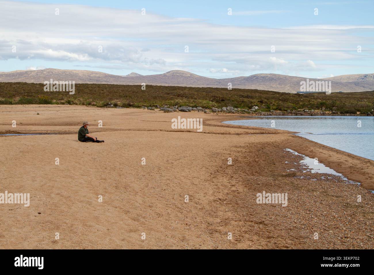 Un uomo seduto su una spiaggia remota tra Loch Laidon e Dubh Lochan alla stazione di Rannoch nelle Highlands scozzesi, con montagne in lontananza. Foto Stock