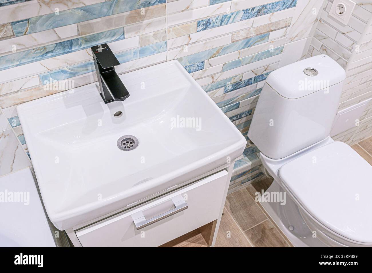 bagno moderno con lavandino bianco con rubinetto nero e wc bianco su una parete piastrellata con dettagli blu e beige. Foto Stock