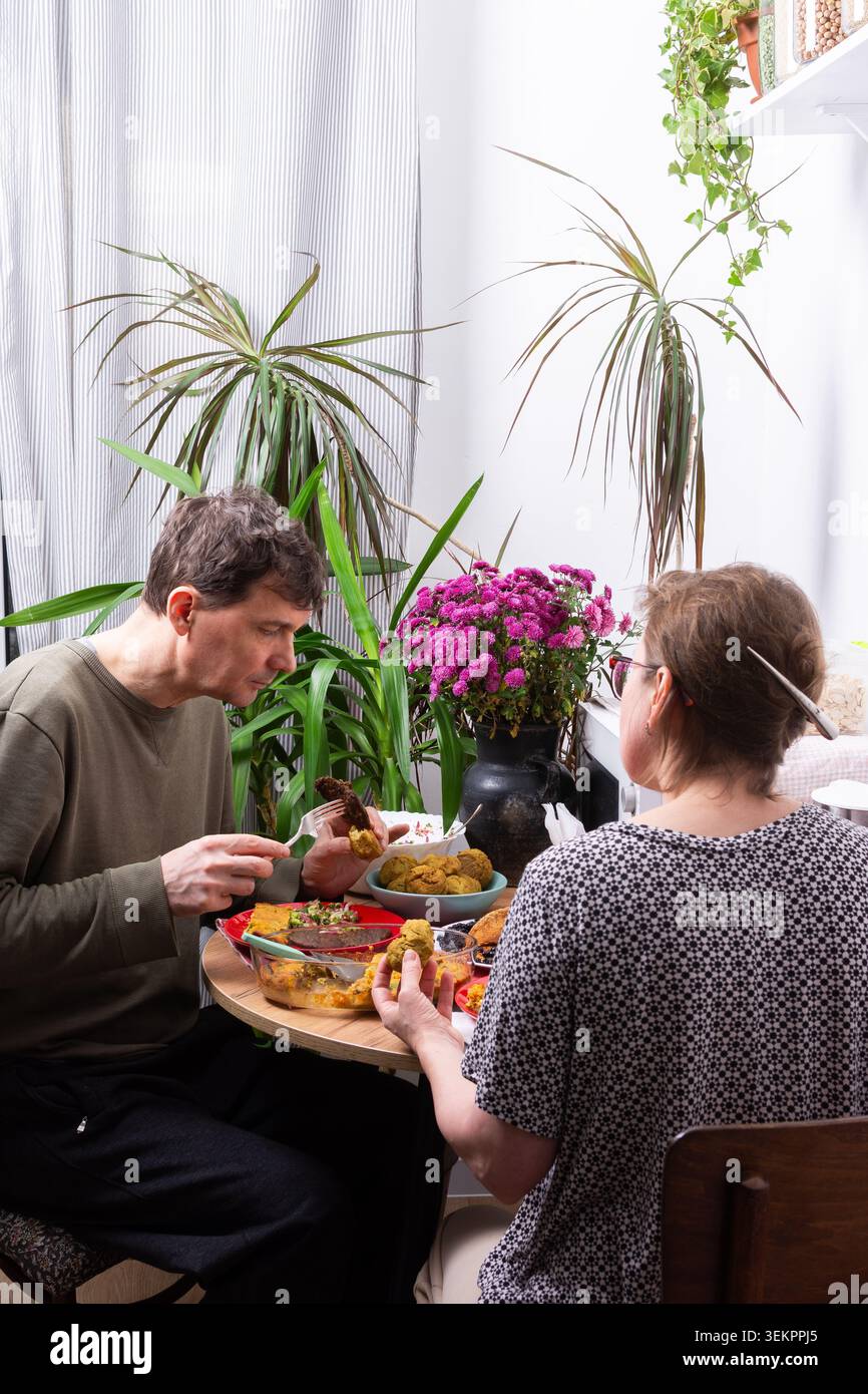 Un uomo e una donna mangiano insieme a un piccolo tavolo. Si siedono vicino a una finestra con piante verdi. I fiori si siedono sul tavolo. Entra la luce del sole, cre Foto Stock