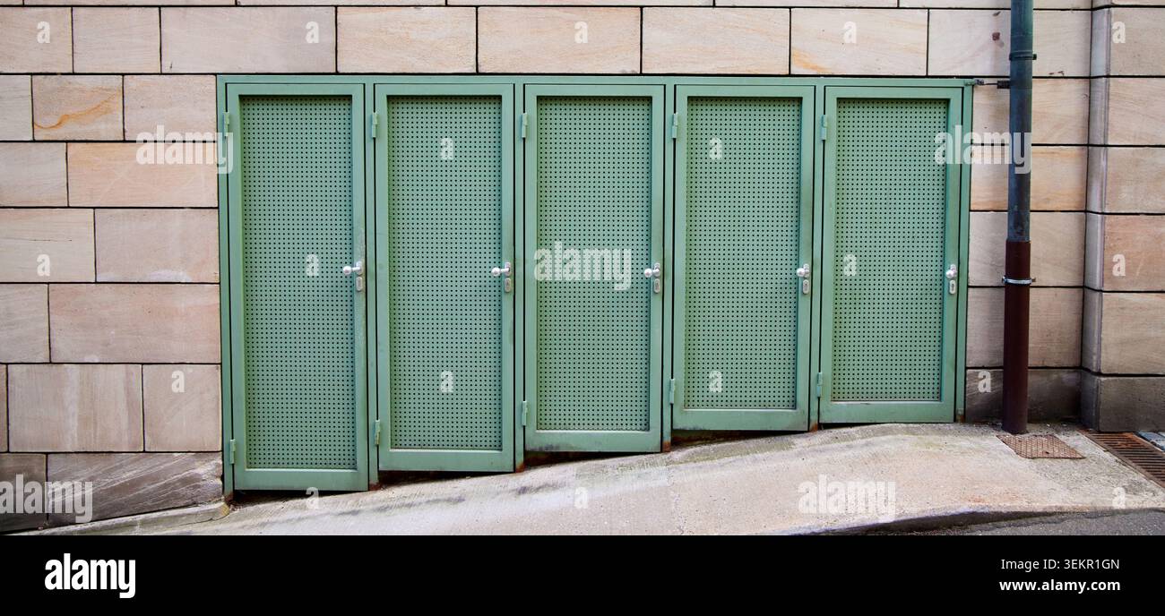 Una fila di cinque porte verdi a rete metallica installate su un pavimento inclinato accanto a un muro di pietra all'aperto. Foto Stock