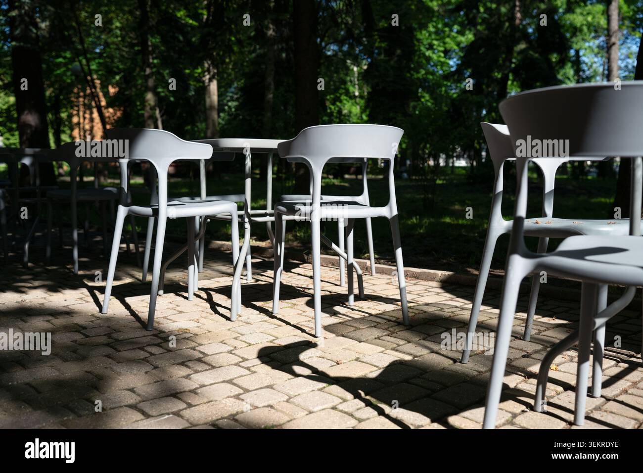 Sedie e tavoli in plastica moderni vuoti su una terrazza pavimentata in un parco estivo. Mobili da caffè all'aperto con ombre a contrasto in una giornata di sole in una zona urbana Foto Stock