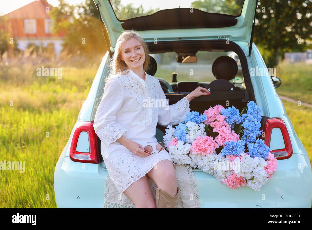 Bella donna con un abito bianco che posa con ortensie colorate in un bagagliaio aperto, creando un'estate da sogno per picnic e vacanze Foto Stock