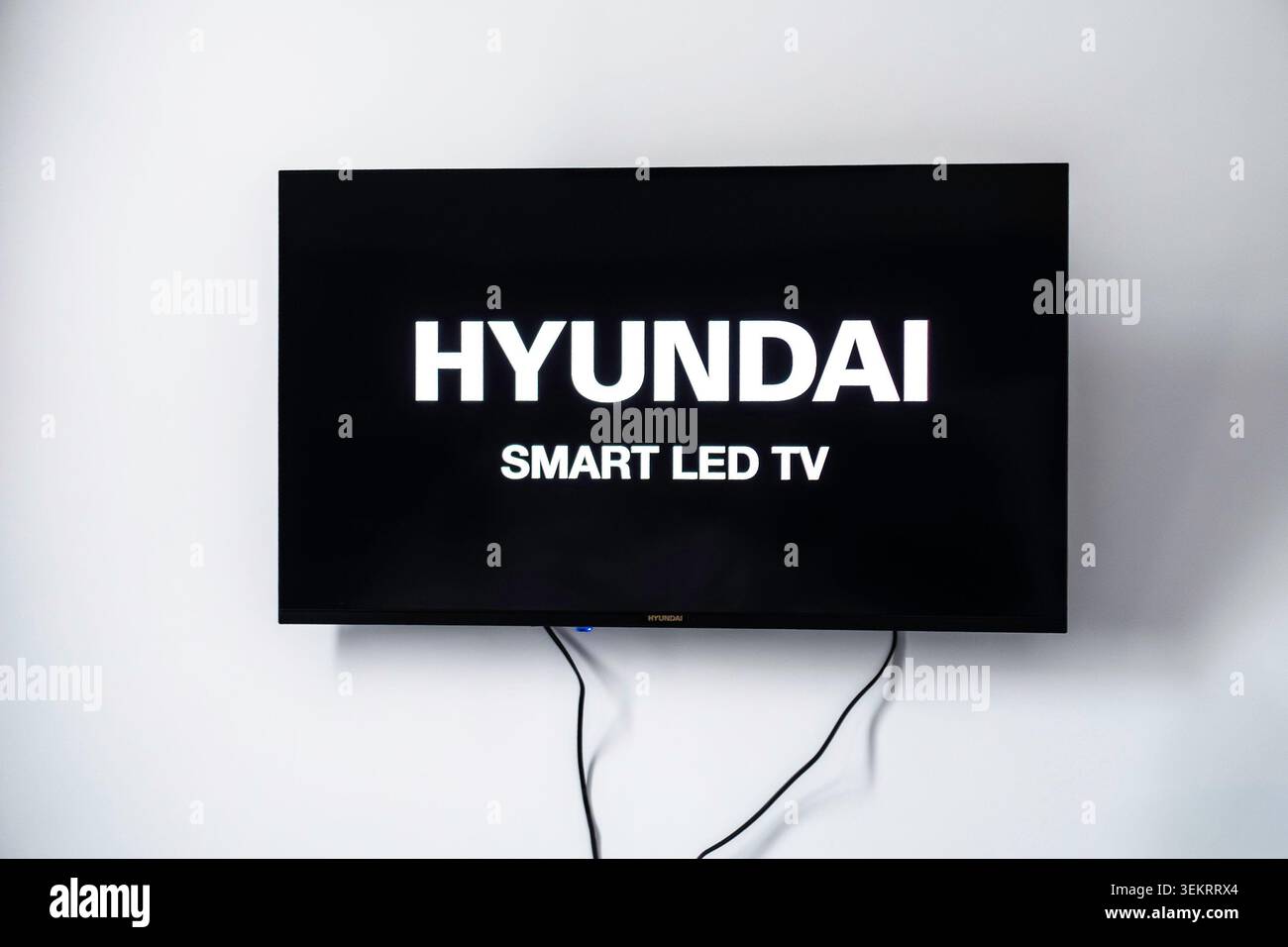 Uno Smart TV LED Hyundai è montato su una semplice parete bianca e mostra la sua schermata di avvio bianca con cavi neri appesi sotto. Foto Stock