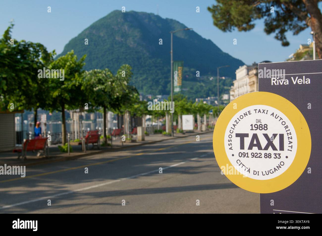 Lugano, Ticino, Svizzera - 24 maggio 2026: Primo piano dell'insegna gialla del servizio taxi ufficiale su Riva Vela. Sullo sfondo del panoramico Monte San Salvatore Foto Stock