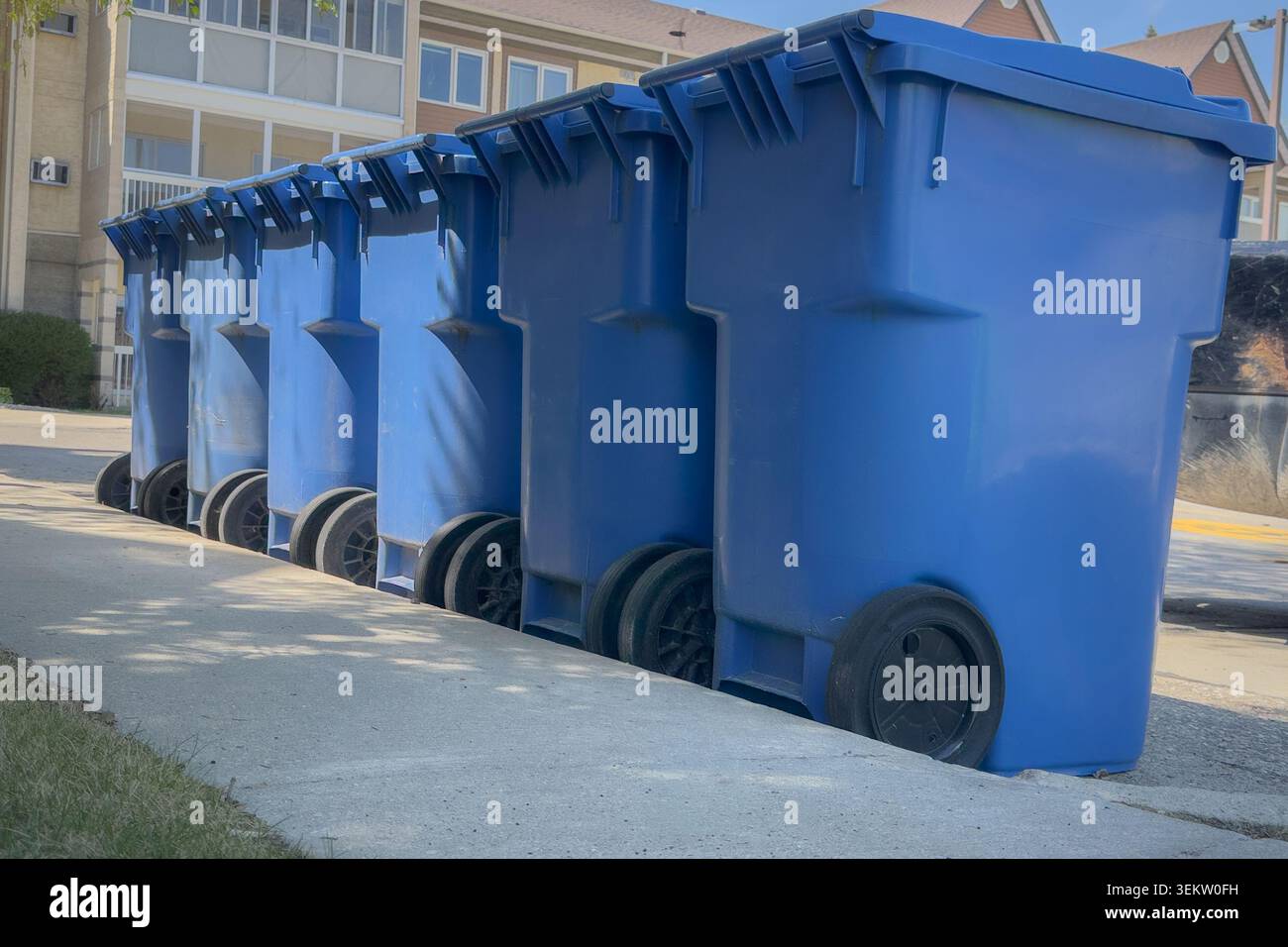 Fila di bidoni di riciclaggio blu collocati lungo una strada residenziale in attesa di raccolta. Concetto di servizio di riciclaggio e gestione dei rifiuti urbani in periferia Foto Stock