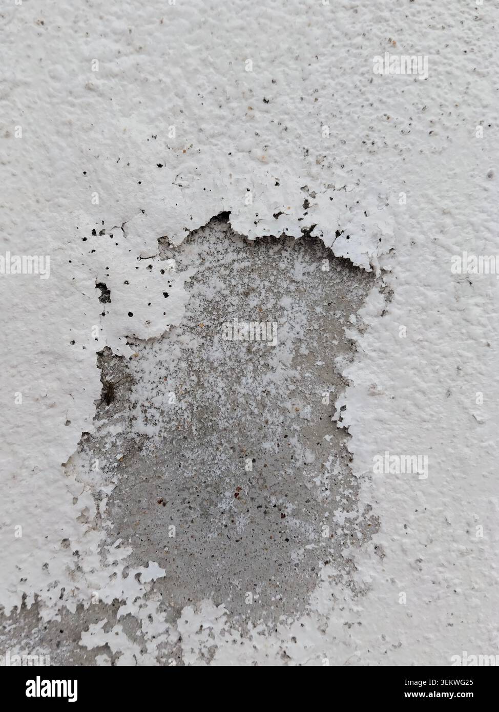 Una vista ravvicinata della vernice bianca sfaldata e scheggiata che si stacca da una parete esterna in cemento, esponendo l'intonaco grezzo e testurizzato grigio sottostante a causa dell'umidità Foto Stock