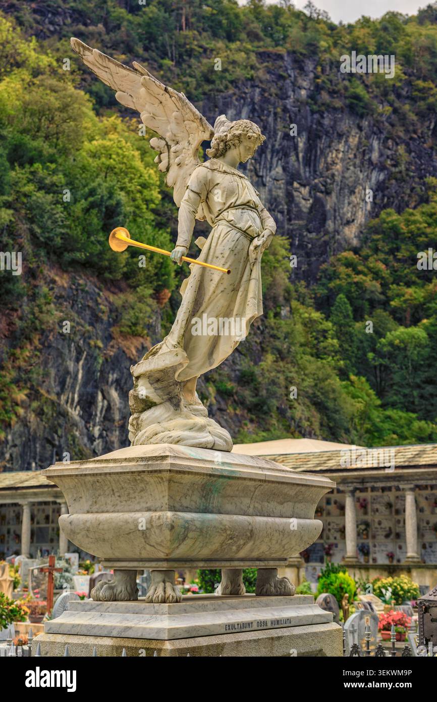 Chiavenna, Italia - 10.10.2022: Figura alata in pietra scolpita con un corno d'oro in un cimitero sostenuto da lussureggiante vegetazione di montagna Foto Stock