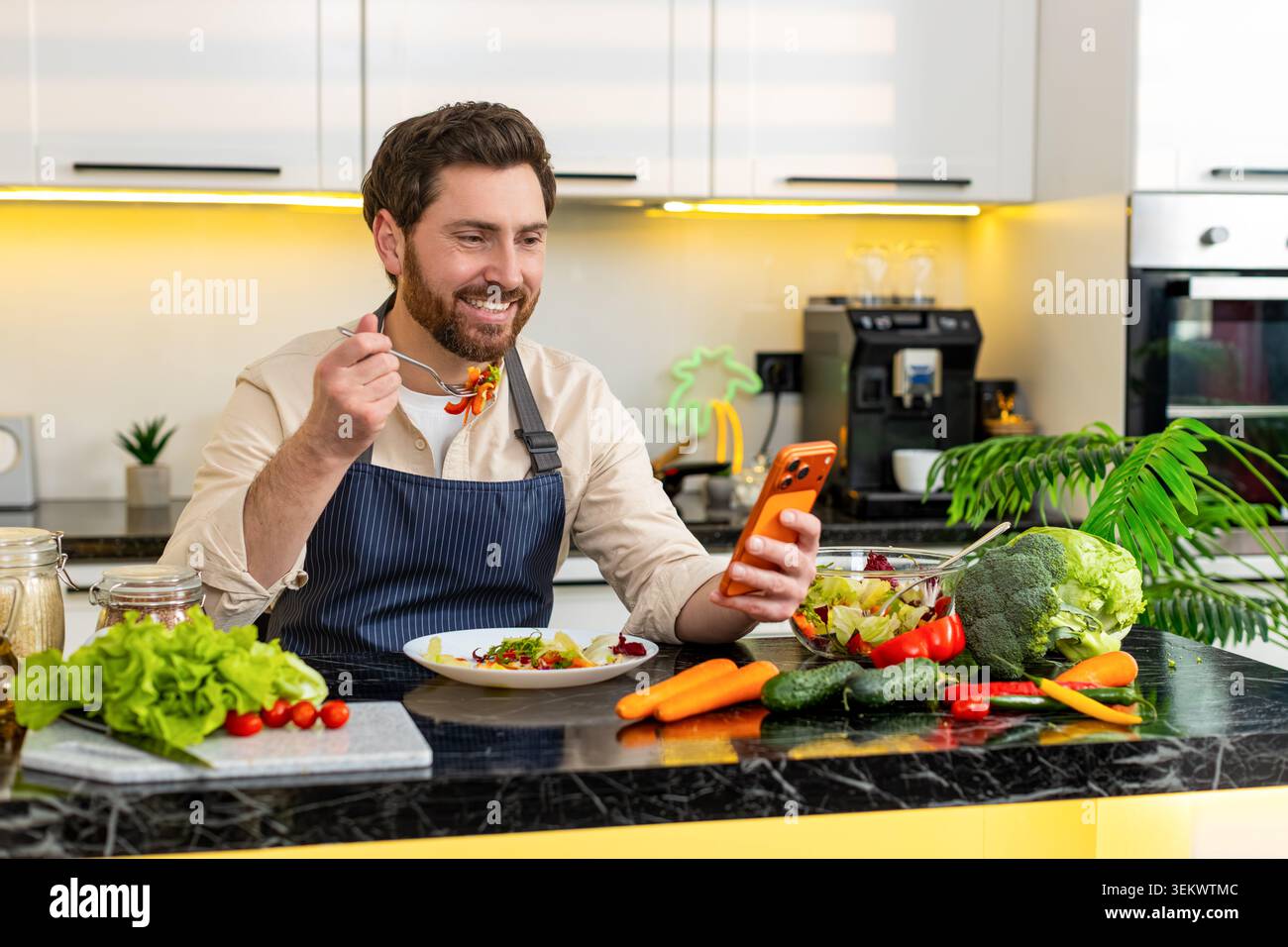Un uomo di mezza età mangia insalata di verdure fresche con sorriso mentre guarda contenuti molto divertenti sullo smartphone nella cucina di casa. Il giovane ragazzo gode di un sano pasto dietetico dopo l'allenamento sentendosi soddisfatto e motivato. Foto Stock