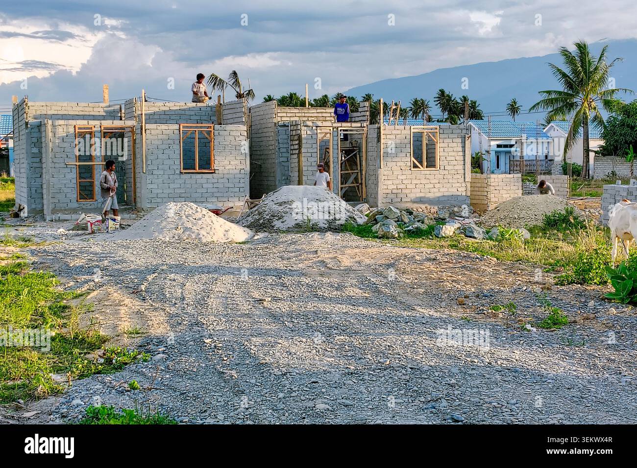 Sulawesi centrale, Indonesia - 21 maggio 2026 : i lavoratori costruiscono una casa su un terreno aperto. I bambini giocano nelle vicinanze, godendosi il sole. Blocchi di cemento e mucchi di sabbia circondano il sito. Le palme sorgono dietro la struttura Foto Stock