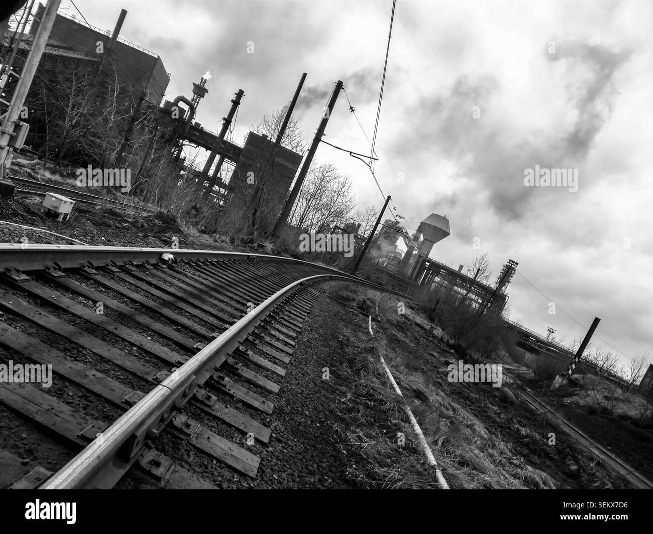 Vista in bianco e nero dei binari ferroviari che conducono attraverso un impianto industriale pesante con cisterne, acciaierie e un cielo nuvoloso Foto Stock