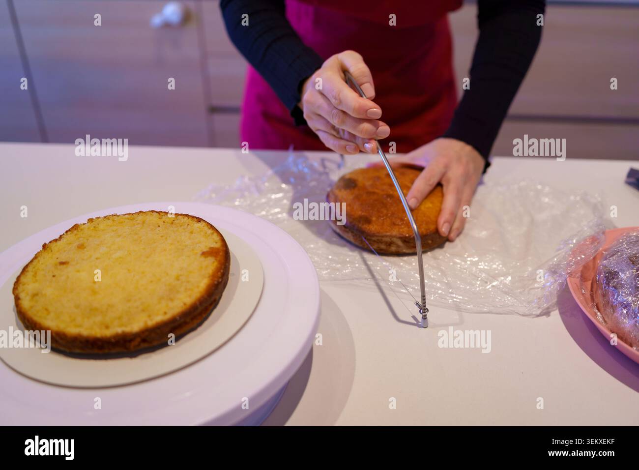 Affettare le mani su uno strato di torta cotto utilizzando un livellatore per torte a filo metallico su un bancone da cucina Foto Stock