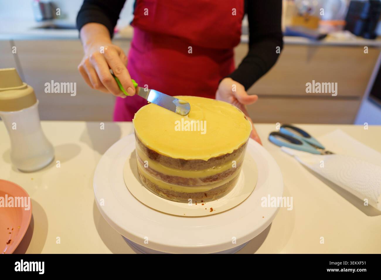 Mani che applicano la glassa cremosa su una torta appena sfornata utilizzando una spatola in cucina Foto Stock