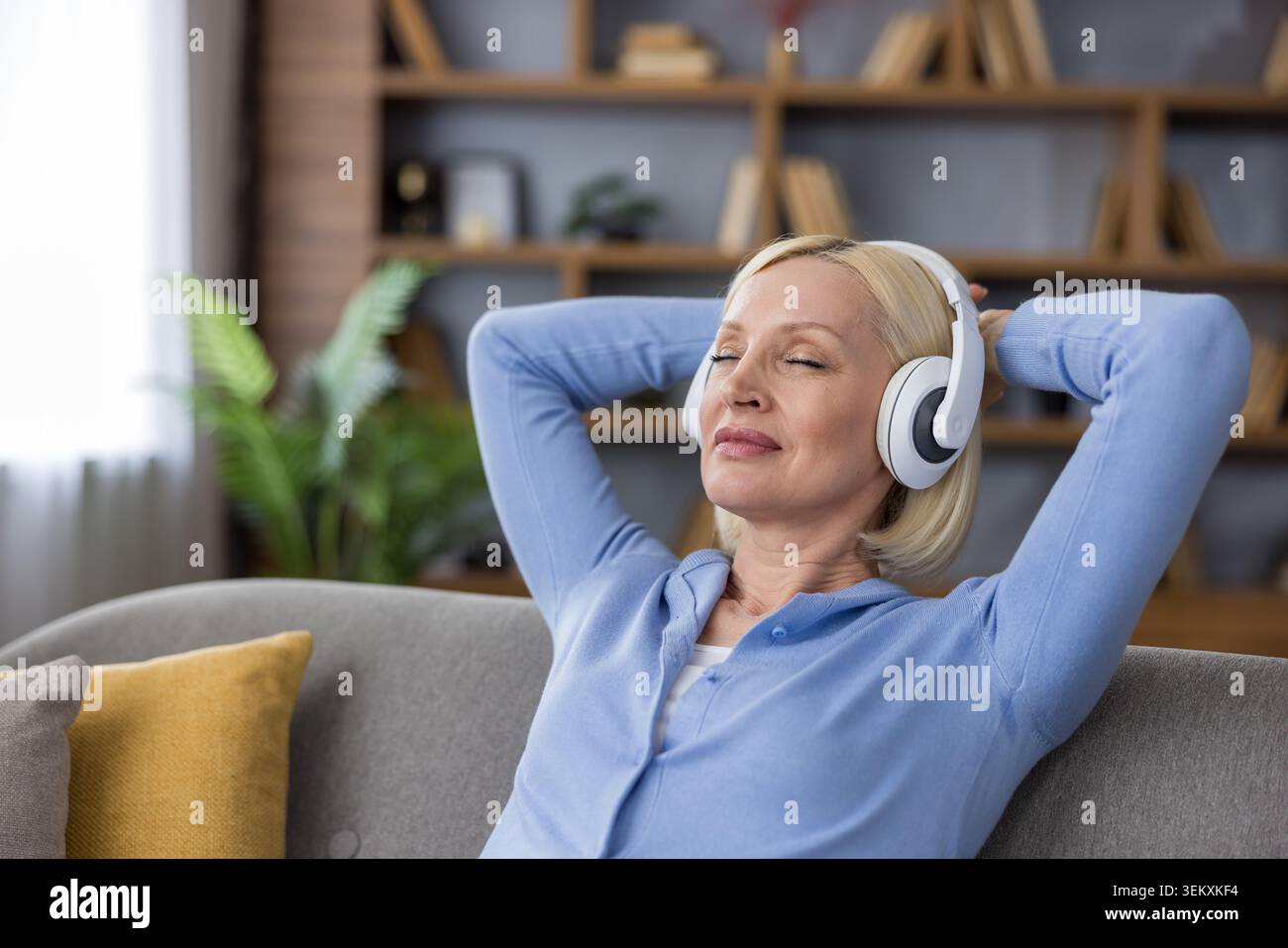 Donna anziana che ascolta la musica con gli occhi chiusi e le cuffie bianche indossate, si rilassa con le mani dietro la testa su un comodo divano nel suo salotto Foto Stock