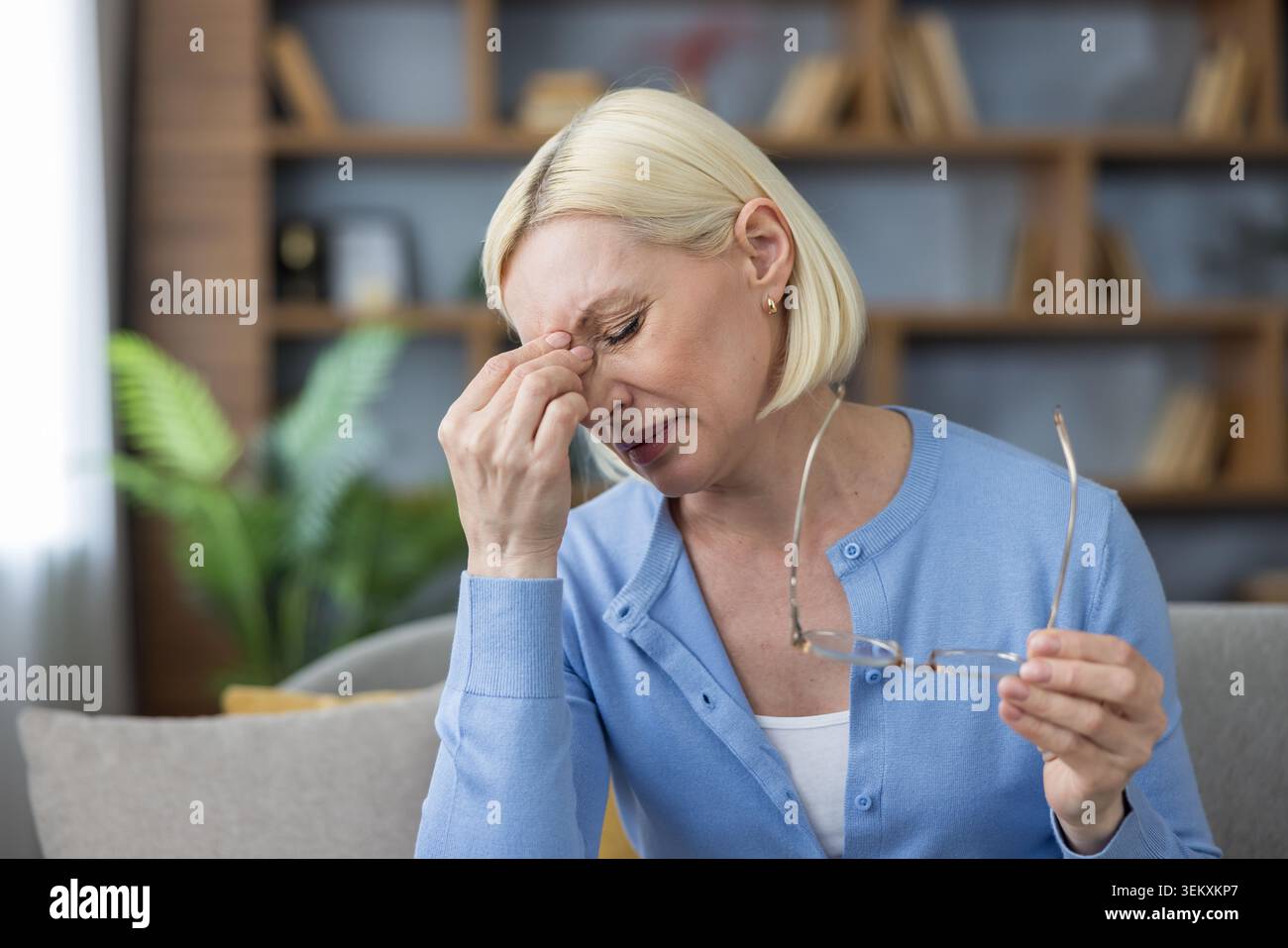 Donna matura con capelli biondi corti che sfregano le stanghette e tengono gli occhiali, affetta da emicrania e affanno gli occhi a casa, stanca, stressata e con dolore e disagio visibili Foto Stock