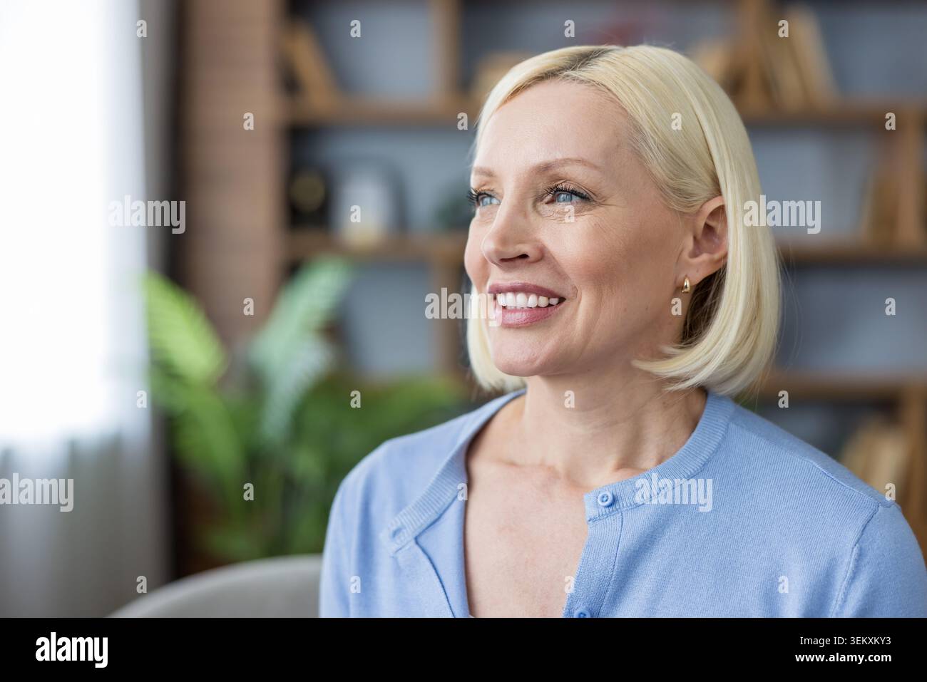 Donna matura con capelli biondi e occhi blu che sorride calorosamente, esprimendo felicità e ottimismo in un ambiente interno confortevole, che rappresenta il benessere e le emozioni positive Foto Stock