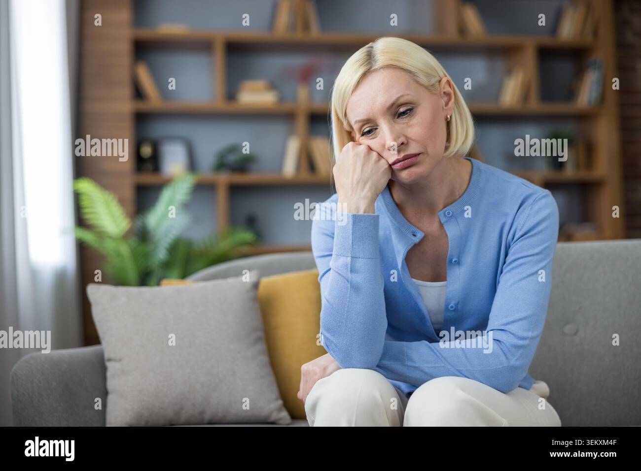 Donna anziana che soffre di malinconia, solitudine e tristezza, soffre di stress emotivo e problemi di salute mentale mentre si siede su un divano nel suo salotto Foto Stock