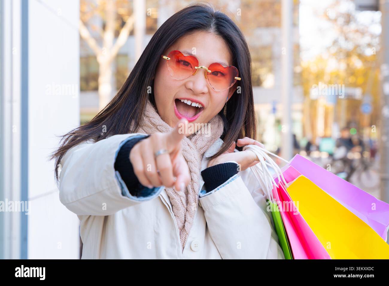 Donna asiatica con occhiali da cuore che puntano alla macchina fotografica, tenendo in mano borse colorate Foto Stock