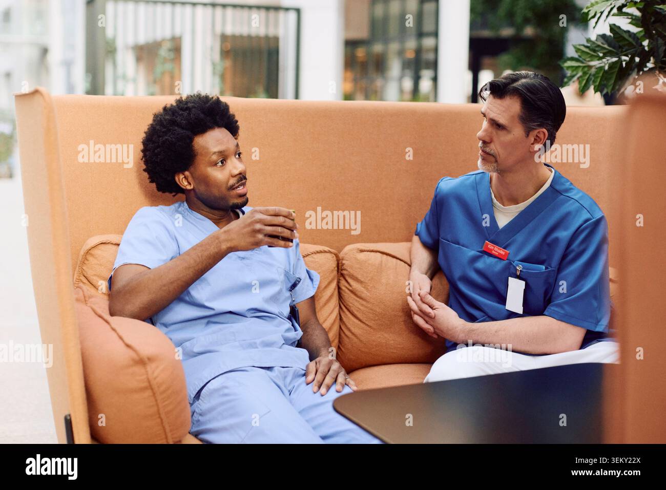 Operatore sanitario maschile che parla con il collega mentre si siede sul divano durante la pausa caffè nella hall dell'ospedale Foto Stock