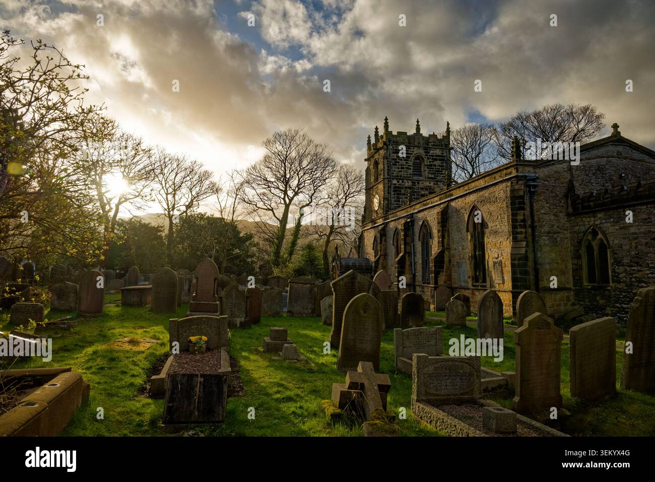 Una chiesa medievale nel suo cimitero con numerose lapidi e alberi bagnati dalla luce invernale serale, Castleton, Peak District, Derbshire. Foto Stock