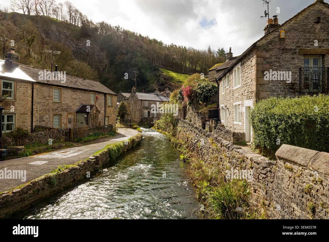 Un piccolo fiume scorre tra cottage in pietra in un villaggio rurale con colline boscose oltre Castleton, Peak District, Derbyshire. Foto Stock