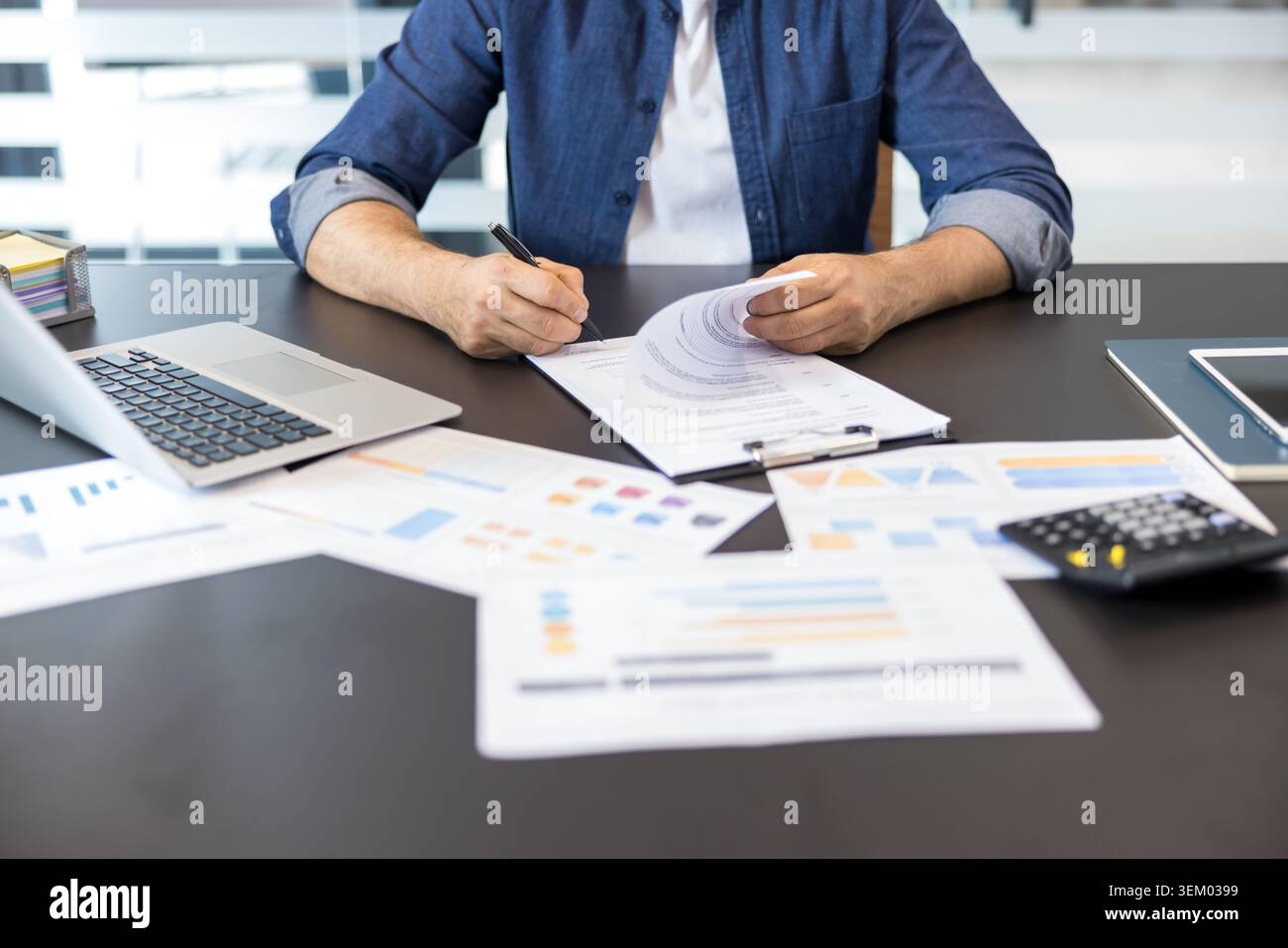 Uomo professionista che firma un contratto aziendale o un documento finanziario presso una moderna scrivania, circondato da grafici analitici, laptop e calcolatrici, gestendo documenti aziendali Foto Stock