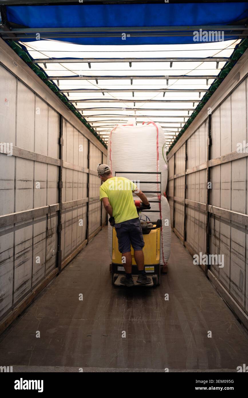 Vista posteriore di un addetto al magazzino caucasico che guida un carrello elevatore a forche elettrico con grandi sacchetti industriali bianchi all'interno di un rimorchio per autocarri. Foto Stock