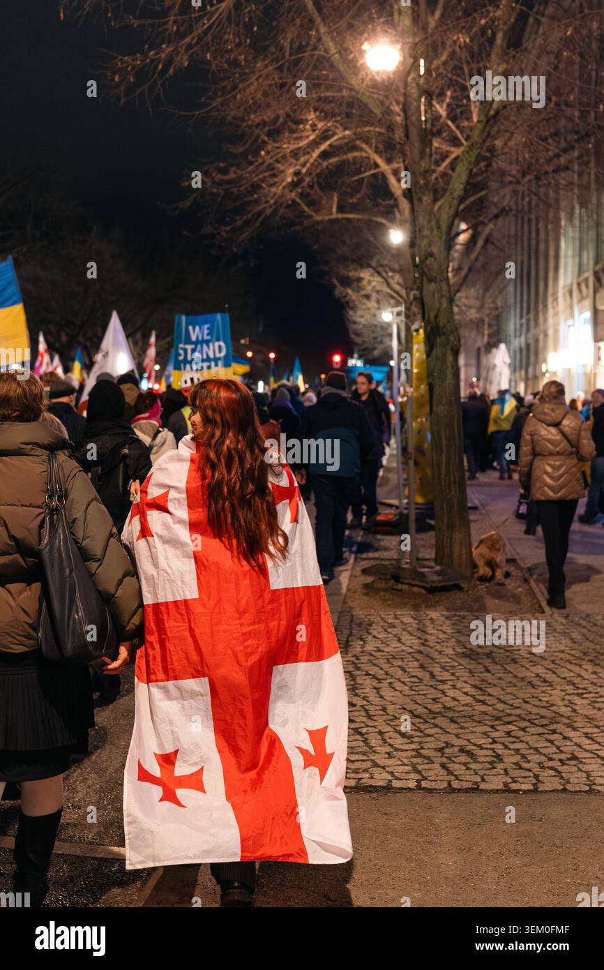 Berlino, Germania - 24 febbraio 2026: Una donna con lunghi capelli cammina lungo un marciapiede della città di notte, drappeggiata nella bandiera nazionale georgiana, mentre si unisce a Foto Stock