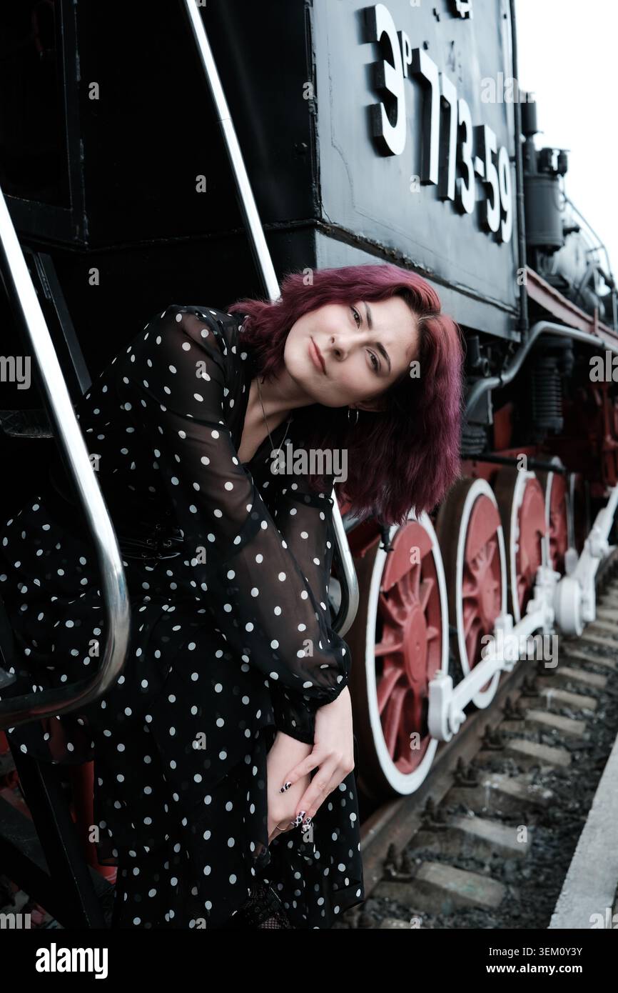Una giovane donna con i capelli viola si siede graziosamente su un classico treno a vapore, nel profondo del pensiero. Foto Stock