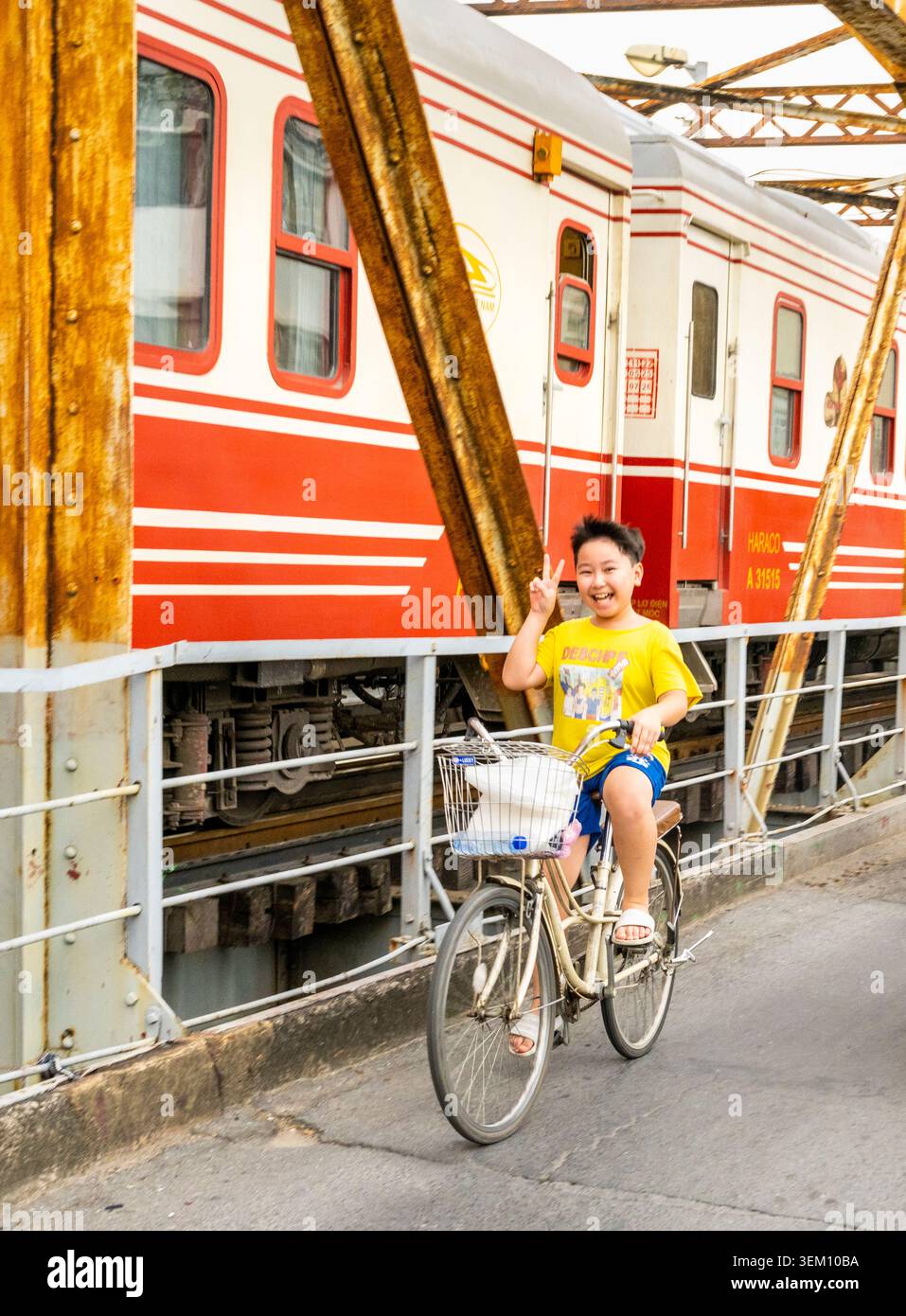 Hanoi, Vietnam-11 giugno 2025: Un treno della Vietnam Railways si avvicina alla stazione ferroviaria di Long Bien, passando davanti a un ragazzo in bicicletta, attraversando il famoso ponte sulla R. Foto Stock