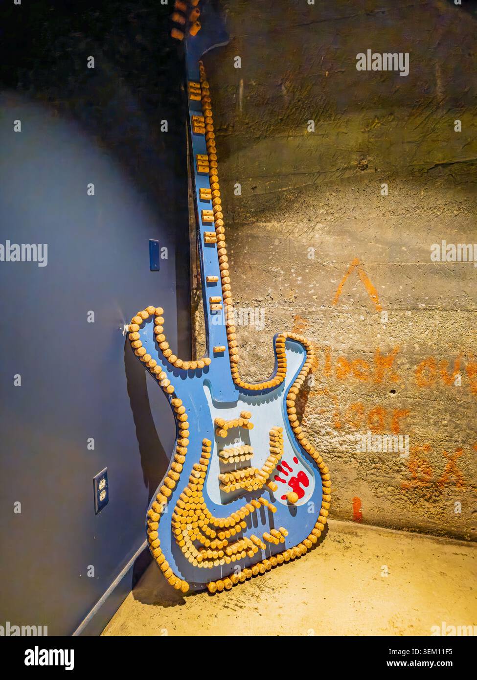 Una grande scultura blu per chitarra elettrica costruita con tappi per bottiglie si appoggia su una parete in cemento con graffiti arancioni. Foto Stock