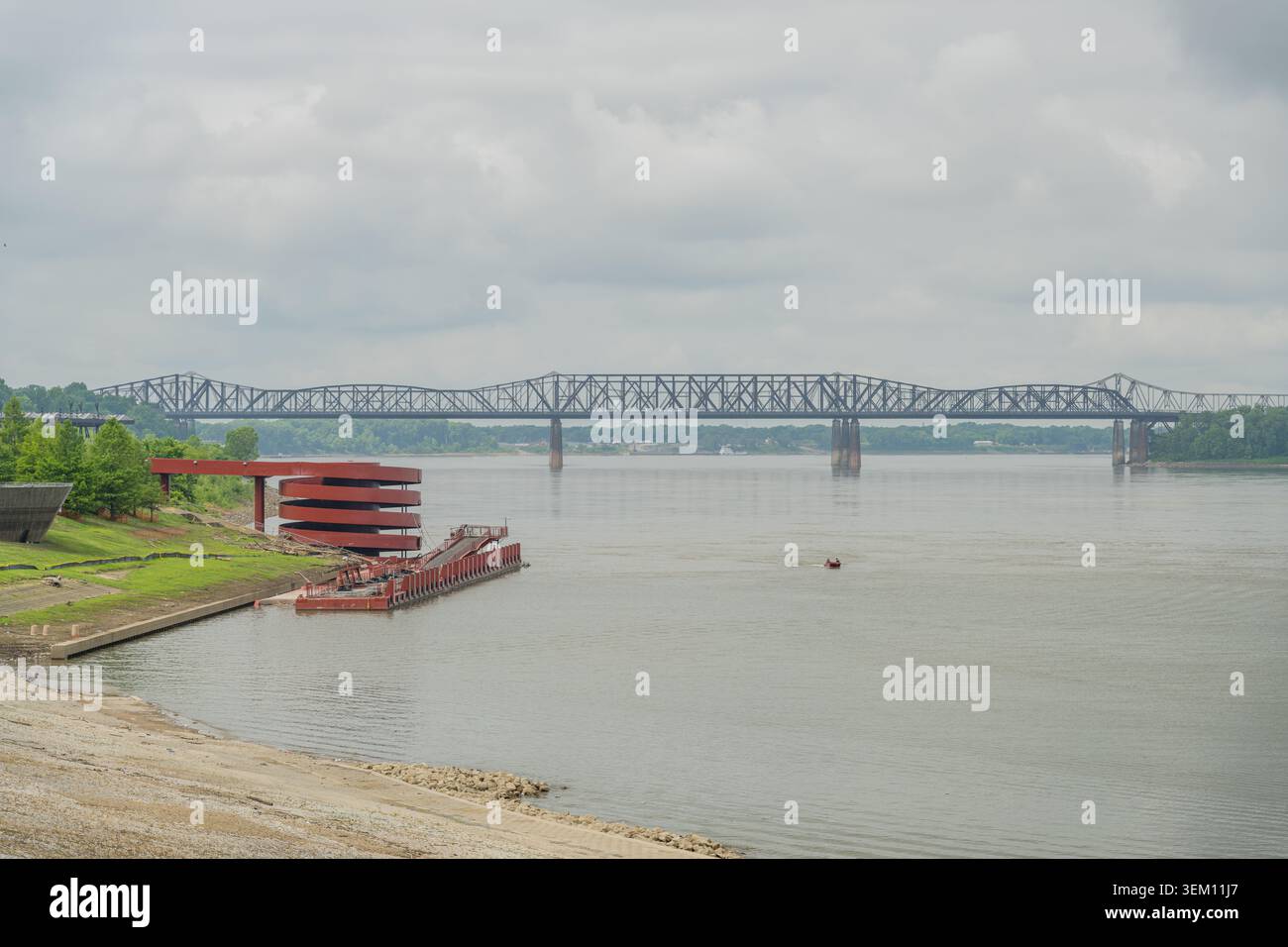Un lungo ponte in acciaio attraversa un ampio fiume sotto un cielo coperto, con una moderna struttura rossa e una rampa per barche sulla riva erbosa. Foto Stock