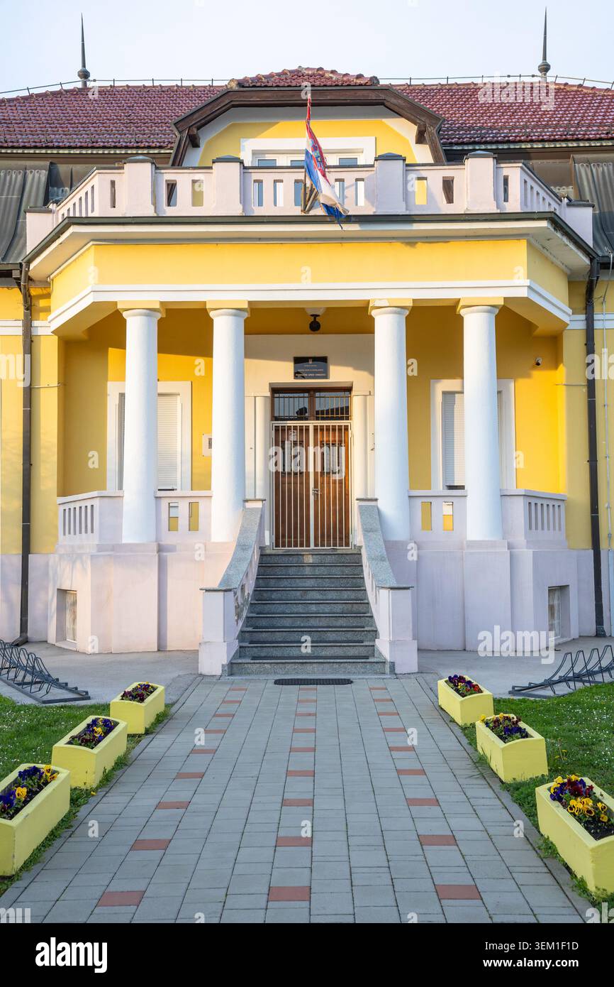 Vinkovci, Croazia - 3 aprile 2026: Elegante villa gialla con colonne classiche, ingresso centrale e tetto in tegole rosse adagiata dietro un passaggio ben curato e. Foto Stock