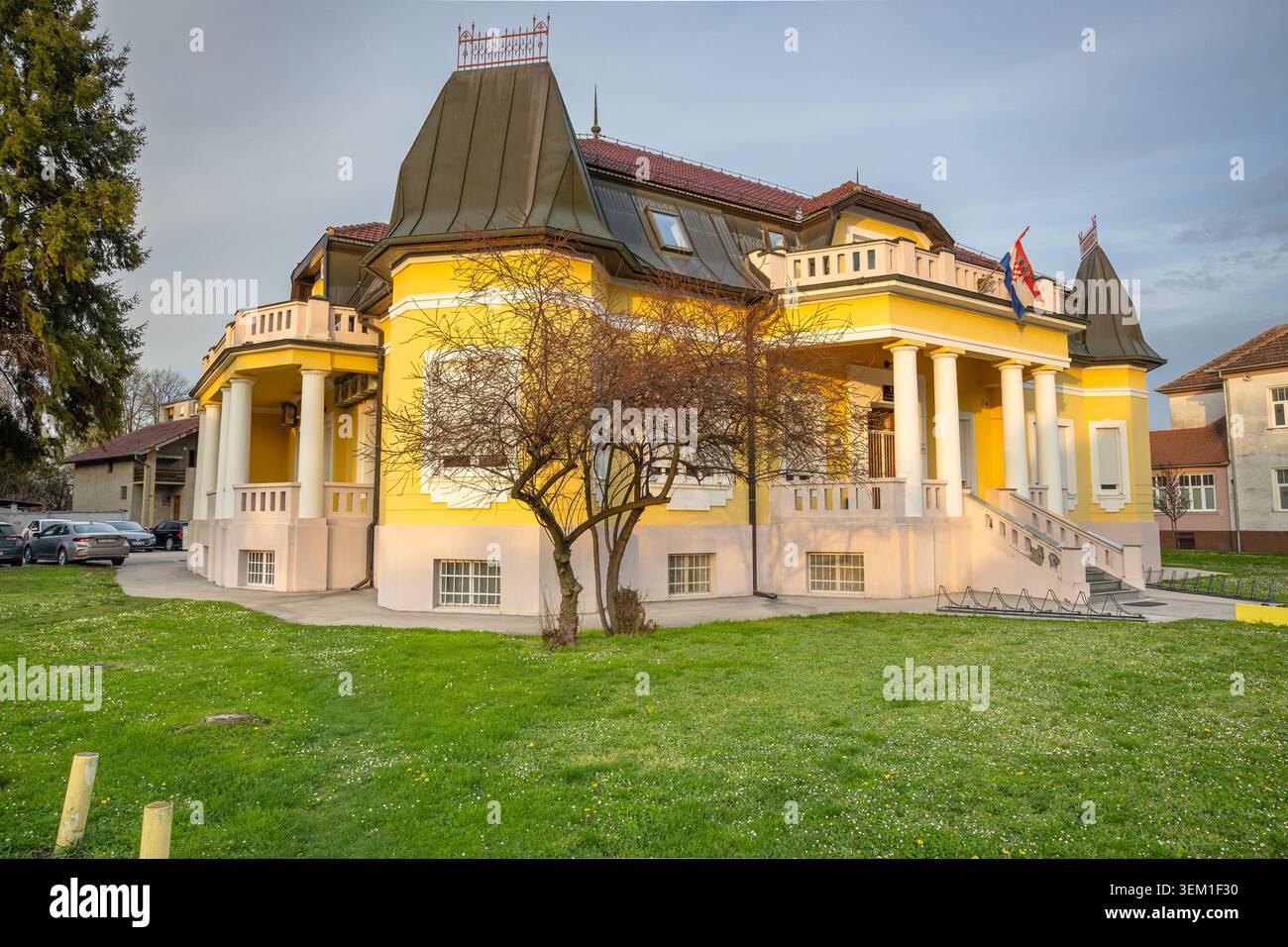Vinkovci, Croazia - 3 aprile 2026: Elegante villa gialla con colonne classiche, ingresso centrale e tetto in tegole rosse adagiata dietro un passaggio ben curato e. Foto Stock