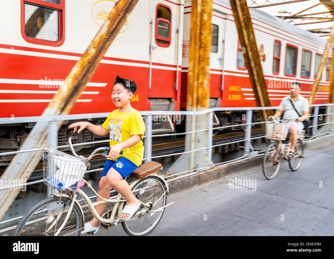 Hanoi, Vietnam-11 giugno 2025: Un treno della Vietnam Railways si avvicina alla stazione ferroviaria di Long Bien, passando davanti a un ragazzo in bicicletta, attraversando il famoso ponte sulla R. Foto Stock