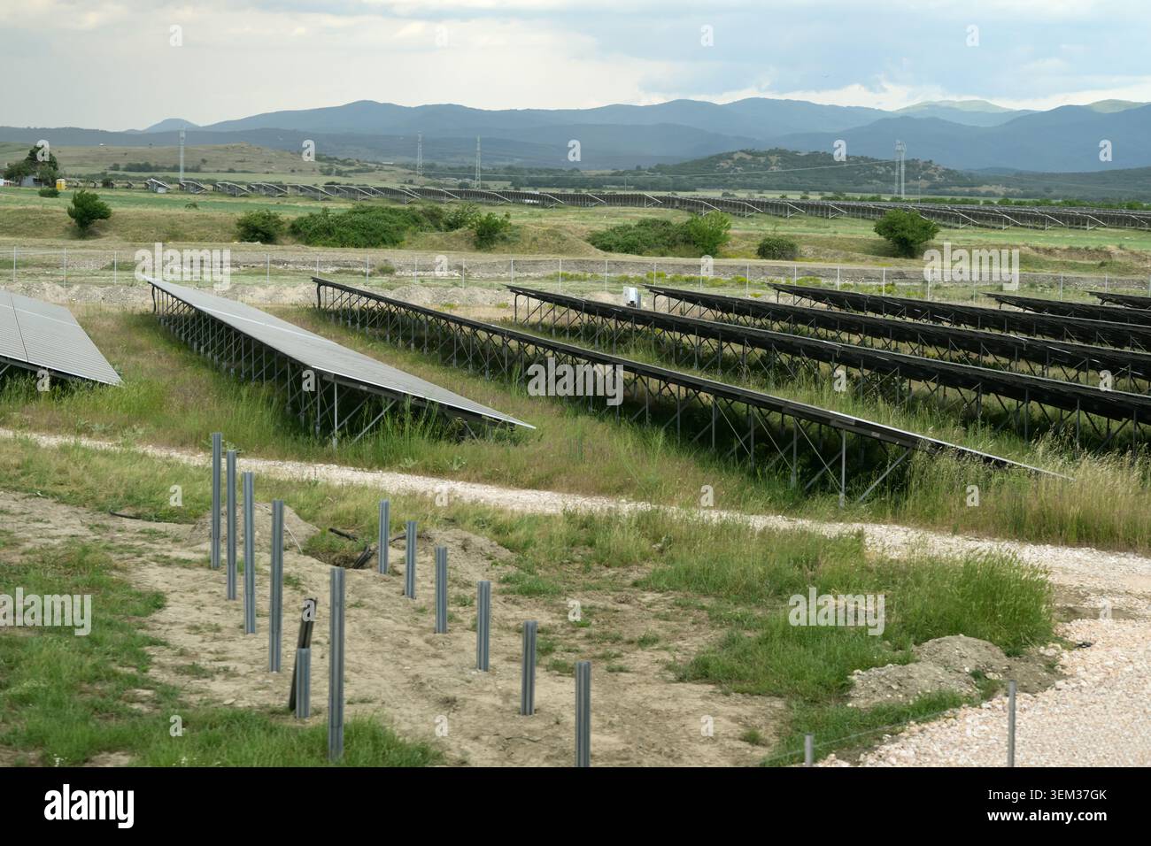 File di pannelli solari e pali metallici appena posizionati mostrano un sito di energia solare in espansione in un paesaggio rurale con colline e cielo nuvoloso. Foto Stock