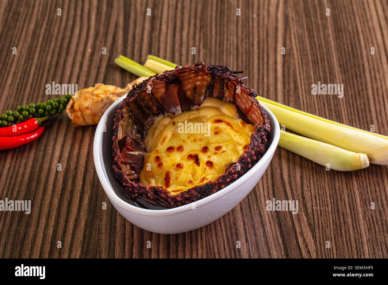 Piatto gourmet di ricci di mare al forno con formaggio fuso e uova in una ciotola bianca, circondato da citronella, peperoncino e grani di pepe su una ta di legno Foto Stock