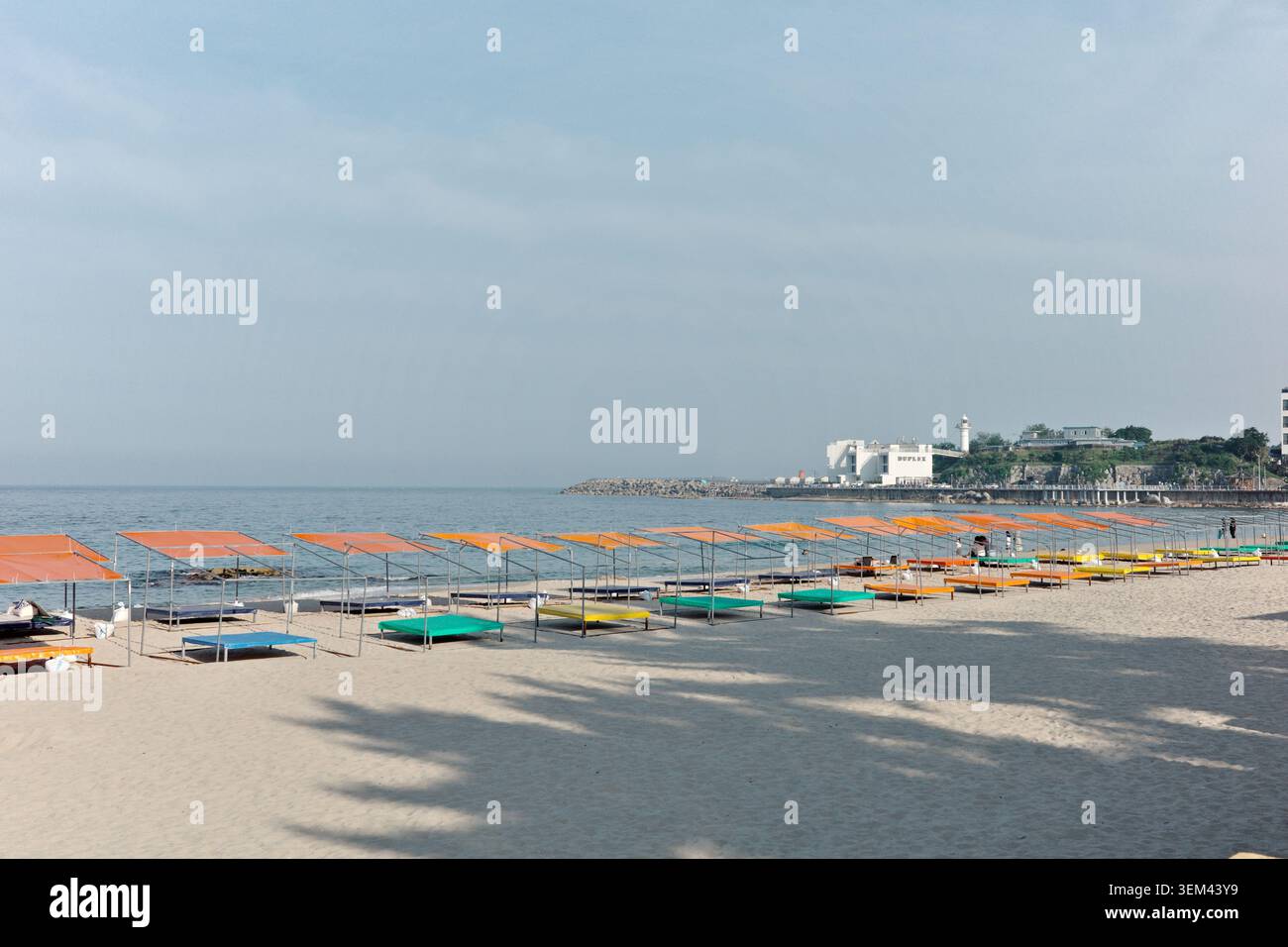 File di colorate ombrelloni da spiaggia con tettoie arancioni e tappeti multicolori su una spiaggia di sabbia bianca con faro e scogliera costiera, Gangneung Foto Stock