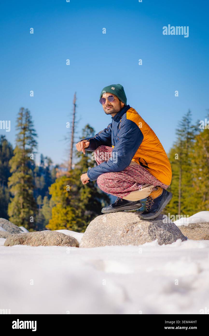 Un giovane uomo che indossa un berretto e una giacca accovacciati su una roccia in un paesaggio innevato di montagna nel Kashmir, India. Foto Stock