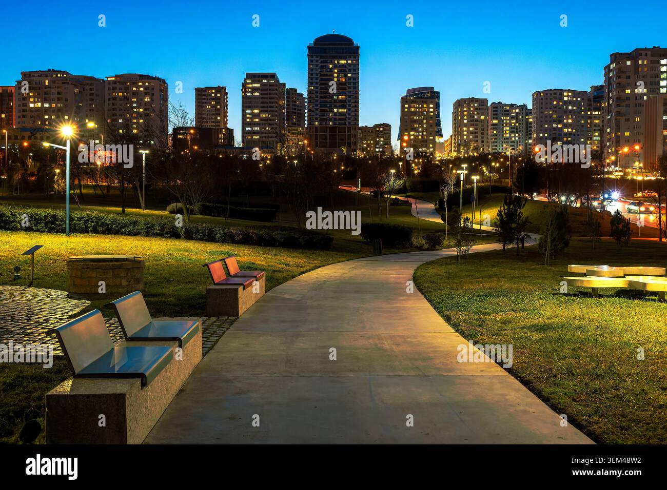 Passerella illuminata attraverso il Parco Mərkəzi con moderne torri residenziali sullo sfondo al tramonto. Baku, Azerbaigian. Foto Stock